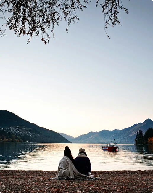 Two people wrapped in blanket on lakeshore watching a boat at sunset