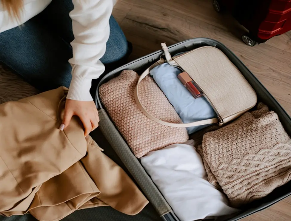 Person packing neutral sweaters and a small crossbody bag into suitcase