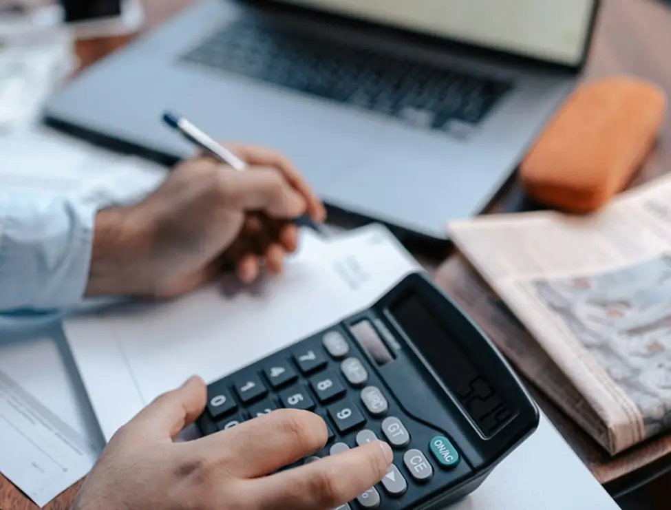 Hands using a calculator and writing on paperwork beside a laptop