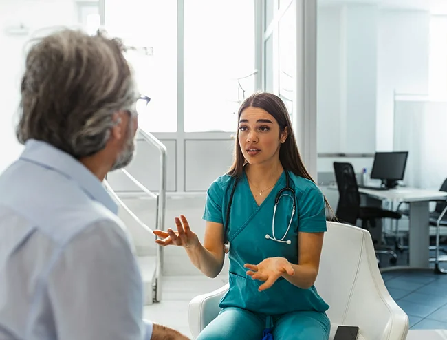 Female nurse in teal scrubs explaining care to older male patient in clinic