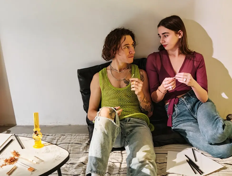 Two people on floor rolling and preparing cannabis with bong